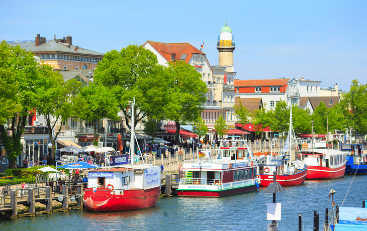 Hafen mit Booten und historischen Gebäuden, Leuchtturm im Hintergrund.
