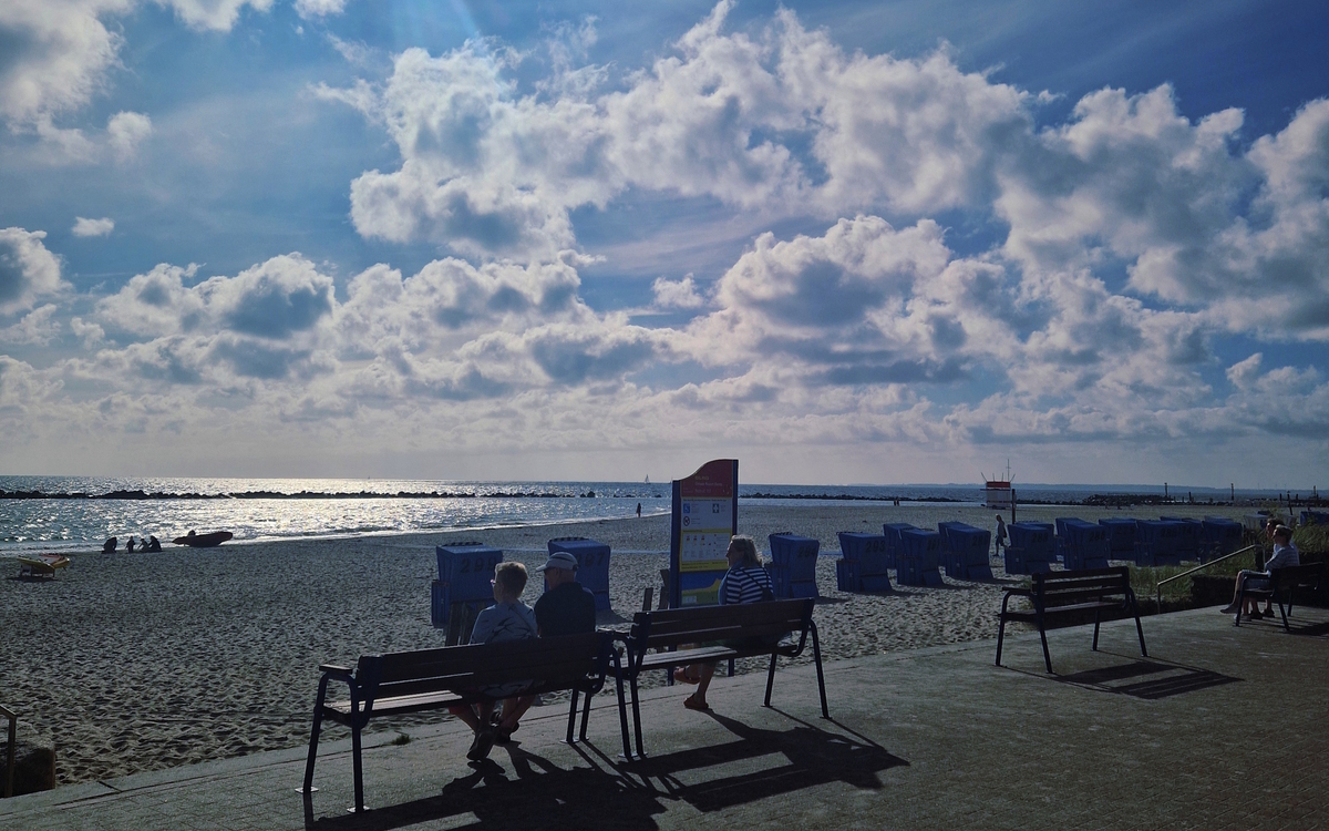 Menschen auf Bänken am Strand von Damp bei Sonnenschein und Wolken am Himmel.