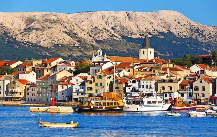 Küstenort mit malerischen Gebäuden, Kirche und Booten im Hafen, im Hintergrund bergige Landschaft.