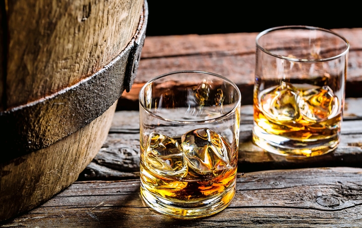 Zwei Whiskygläser mit Eis auf einem Holztisch neben einem Fass.
