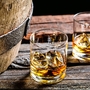Zwei Whiskygläser mit Eis auf einem Holztisch neben einem Fass.
