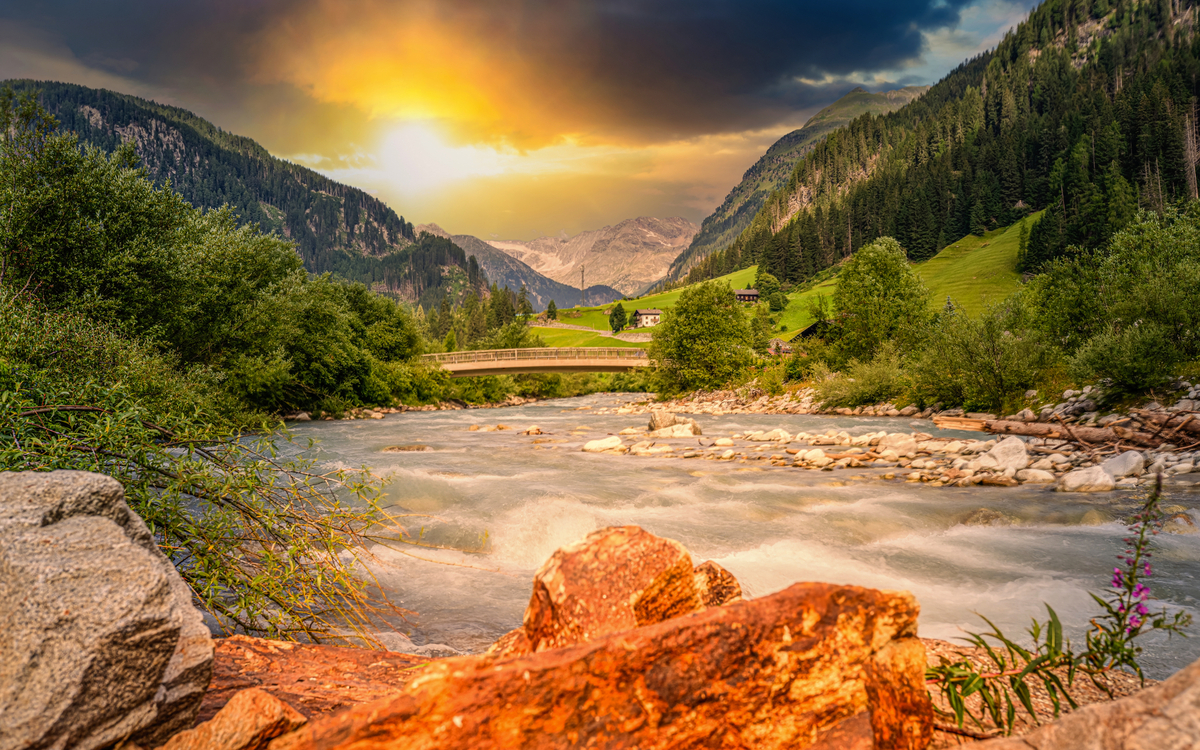 Fluss in Gebirgslandschaft bei Sonnenuntergang, umgeben von Bäumen und Bergen.