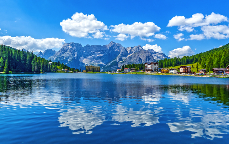Bergsee mit umliegenden Bergen, Wald und Häusern bei blauem Himmel und Wolken.
