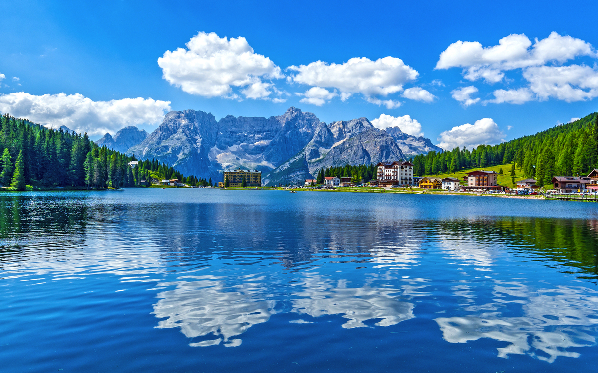 Bergsee mit umliegenden Bergen, Wald und Häusern bei blauem Himmel und Wolken.
