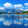 Bergsee mit umliegenden Bergen, Wald und Häusern bei blauem Himmel und Wolken.