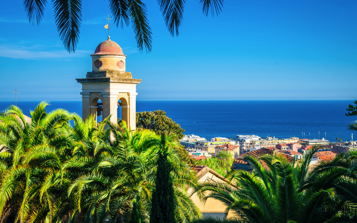 Glockenturm vor der Kulisse von Sanremo an der ligurischen Küste mit Blick auf das Meer und Palmen.