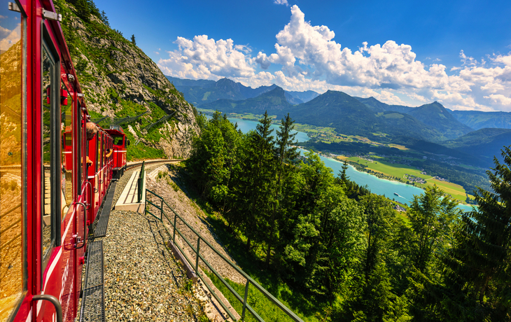 Rote Zahnradbahn fährt am Berg, Panorama mit See und Bergen im Hintergrund.
