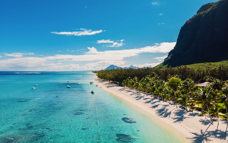 Strand von Mauritius mit türkisfarbenem Wasser, Palmen und einer Bergkulisse im Hintergrund.