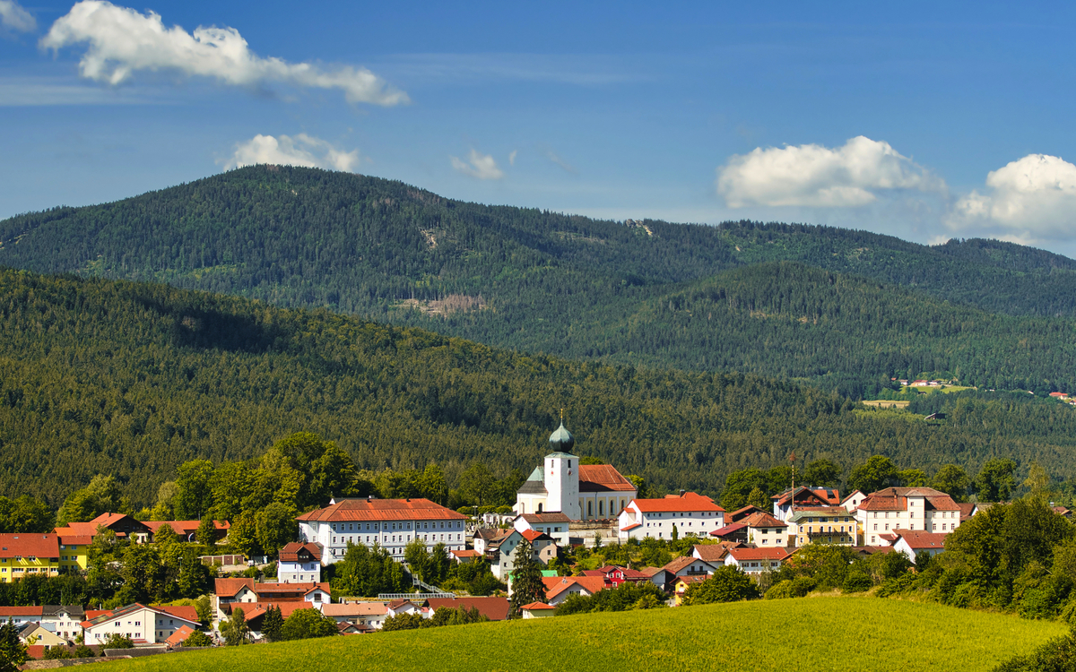 Kleinstadt in hügeliger Landschaft mit Kirche, Wälder im Hintergrund, blauer Himmel.