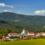 Kleinstadt in hügeliger Landschaft mit Kirche, Wälder im Hintergrund, blauer Himmel.