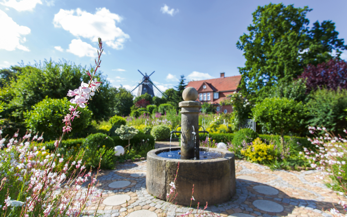 Brunnen im Park mit Mühle und blühenden Blumen im Hintergrund.