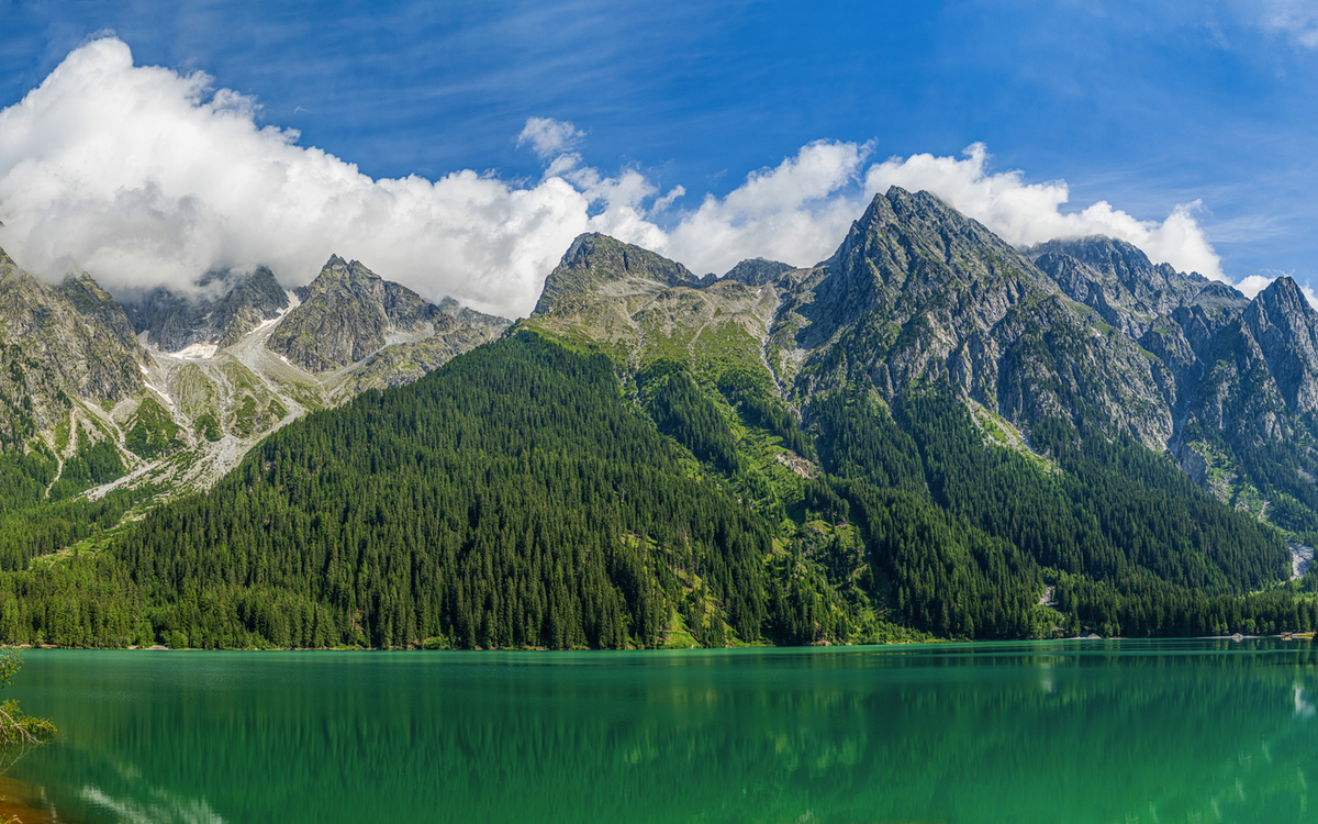 Bergsee mit grüner Wasserfläche und bewaldeten Bergen im Hintergrund bei klarem Himmel.