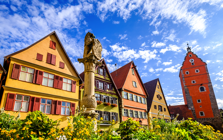 Bunte Fachwerkhäuser und ein Brunnen in einer Altstadt bei blauem Himmel.