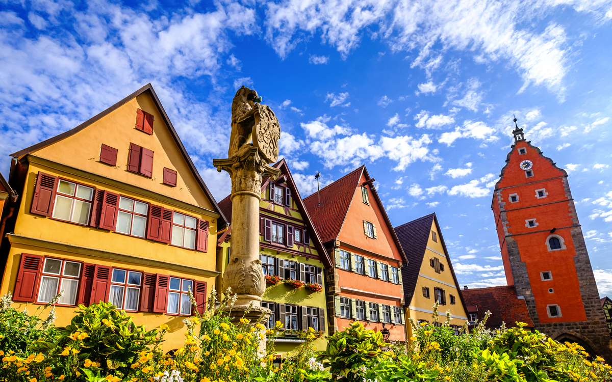 Bunte Fachwerkhäuser und ein Brunnen in einer Altstadt bei blauem Himmel.