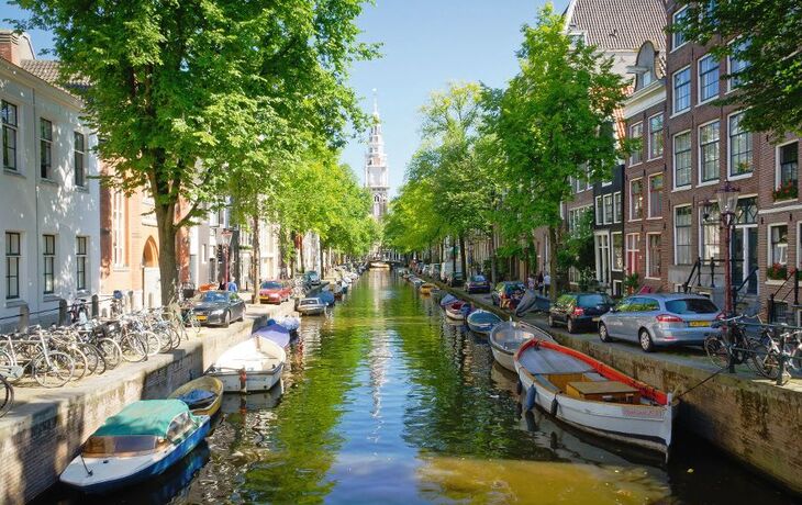 Blick auf eine typische Amsterdamer Gracht mit Booten, gesäumt von Bäumen und traditionellen niederländischen Gebäuden.