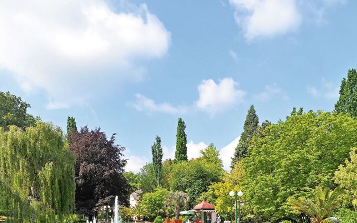 Ein Park mit grünen Bäumen, Springbrunnen und Sitzbänken unter blauem Himmel.