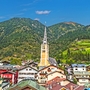 Kirche mit spitzem Turm vor grünen bewaldeten Bergen und bunten Häusern im Vordergrund.