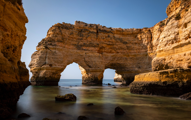 Felsbogen an der Algarve-Küste bei Sonnenuntergang, ruhiges Meer im Vordergrund.
