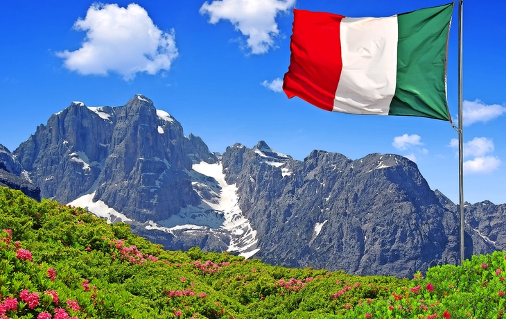 Italienische Flagge weht vor Bergkulisse mit Blumenwiese.