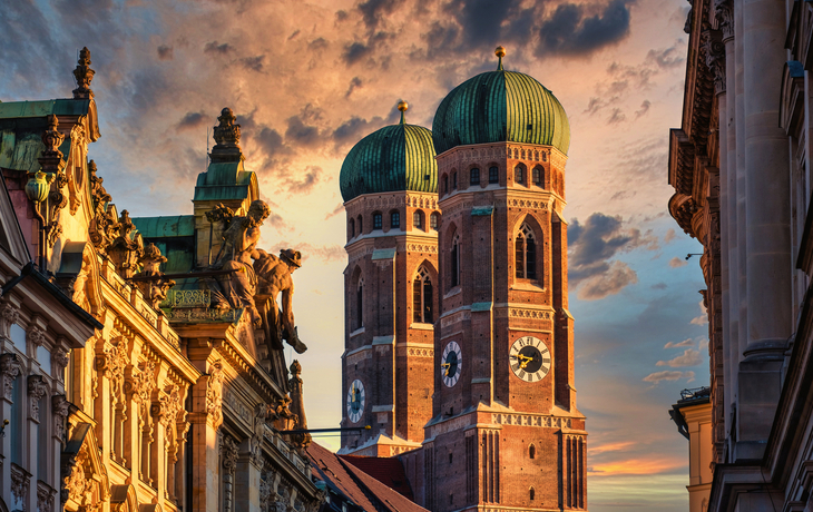 Frauenkirche in München bei Sonnenuntergang mit malerischem Himmel und Gebäudefassaden im Vordergrund.