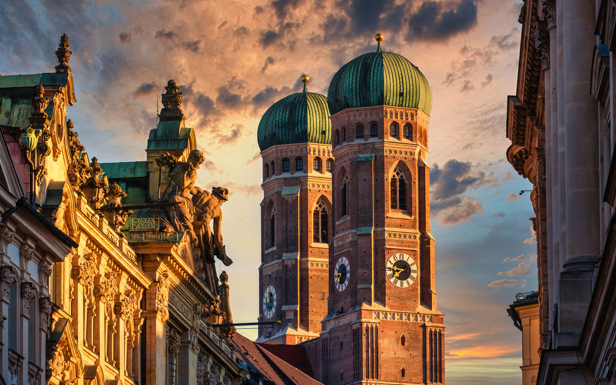 Frauenkirche in München bei Sonnenuntergang mit malerischem Himmel und Gebäudefassaden im Vordergrund.