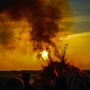 Menschenmenge beobachtet ein großes Lagerfeuer bei Sonnenuntergang, aus dem Rauch aufsteigt.
