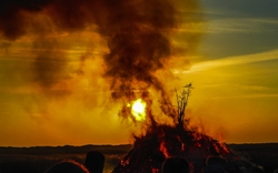 Menschenmenge beobachtet ein großes Lagerfeuer bei Sonnenuntergang, aus dem Rauch aufsteigt.