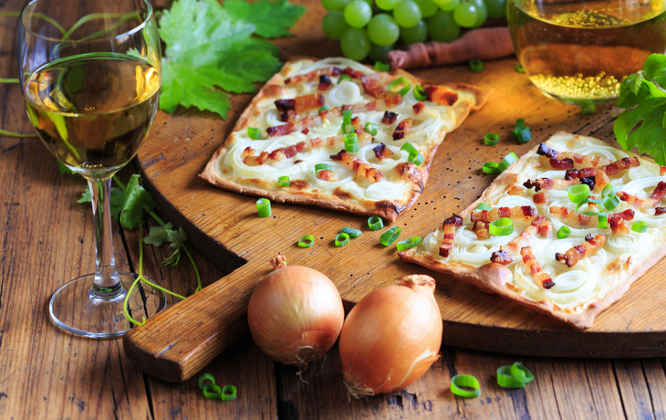 Flammkuchen mit Speck und Zwiebeln auf Holzbrett, daneben Weißwein und Trauben.