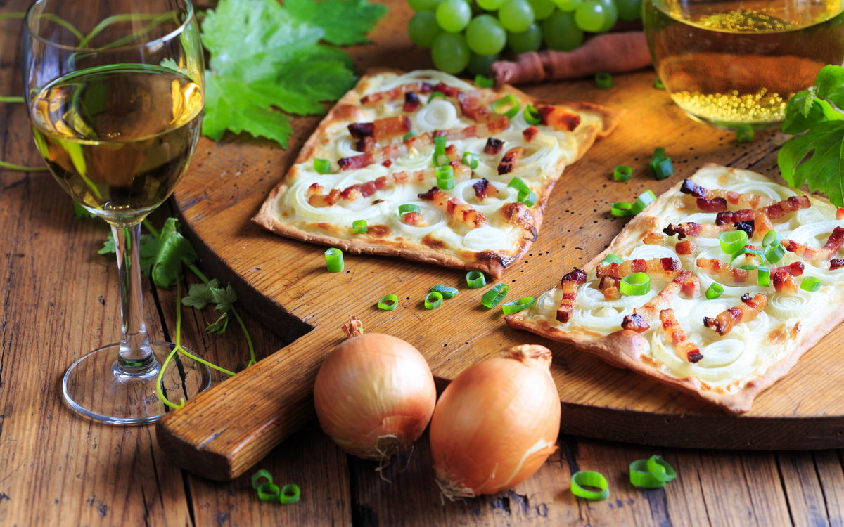 Flammkuchen mit Speck und Zwiebeln auf Holzbrett, daneben Weißwein und Trauben.