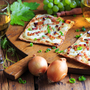 Flammkuchen mit Speck und Zwiebeln auf Holzbrett, daneben Weißwein und Trauben.