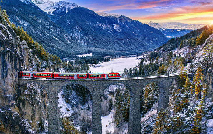 Zug auf Viadukt in winterlicher Berglandschaft bei Sonnenuntergang.