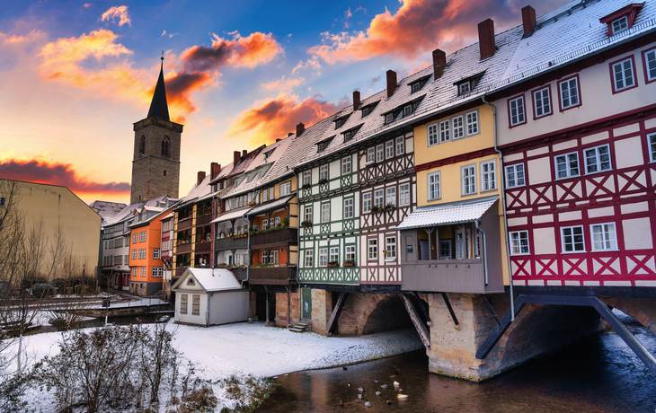 Winterliche Brücke mit Fachwerkhäusern und Kirchturm im Hintergrund bei Sonnenuntergang.