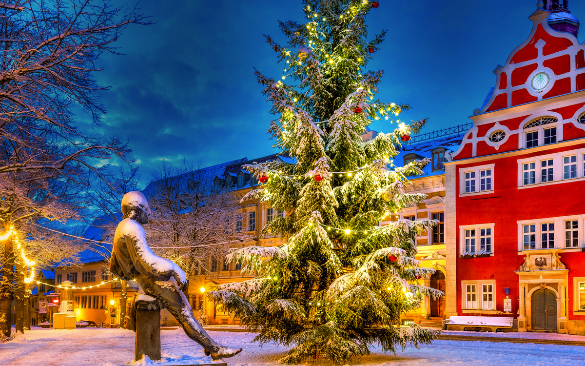 Verschneiter Platz mit Weihnachtsbaum und rotem Gebäude im Hintergrund.