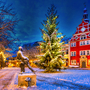 Verschneiter Platz mit Weihnachtsbaum und rotem Gebäude im Hintergrund.