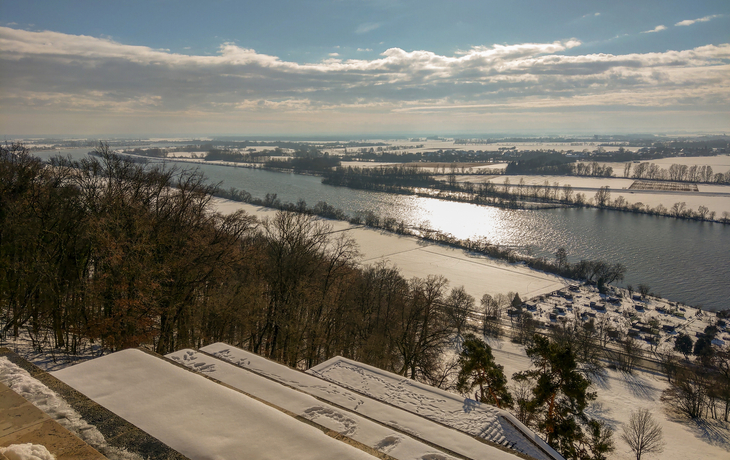 Blick von der Gedenkstätte Walhalla in Donaustauf, Bayern Winterlandschaft mit verschneitem Flussufer und Bäumen unter blauem Himmel.