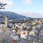 Verschneite Stadt mit Kirchturm und Hügeln im Hintergrund.