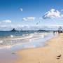 Strandpromenade mit Möwen und Menschen, Holzsteg im Hintergrund bei blauem Himmel.