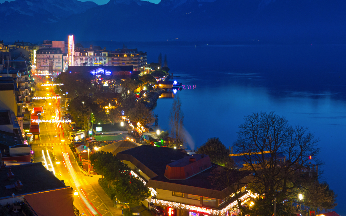 Stadtansicht bei Nacht mit beleuchtetem Ufer und Bergen im Hintergrund.