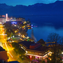 Stadtansicht bei Nacht mit beleuchtetem Ufer und Bergen im Hintergrund.