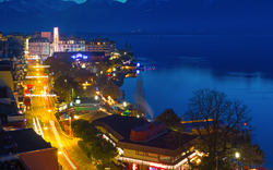 Stadtansicht bei Nacht mit beleuchtetem Ufer und Bergen im Hintergrund.