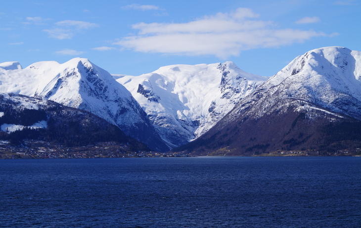 Verschneite Berge am Rand eines ruhigen Sees unter blauem Himmel.