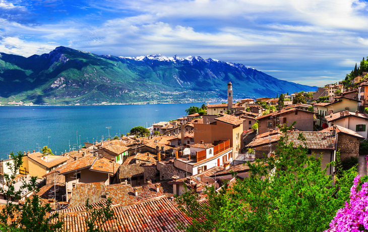 Panoramablick auf Limone sul Garda am Gardasee mit malerischen Häusern und bunten Blumen im Vordergrund, umgeben von Bergen und dem blauen Wasser des Sees.