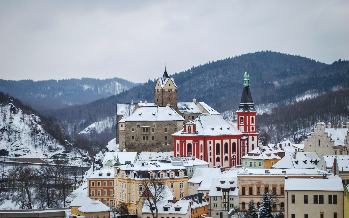 Loket Verschneite Stadt mit Kirche und Burg vor bewaldeten Hügeln im Hintergrund.