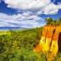 Bunte Sandsteinfelsen vor bewaldeter Landschaft und blauem Himmel mit Wolken.