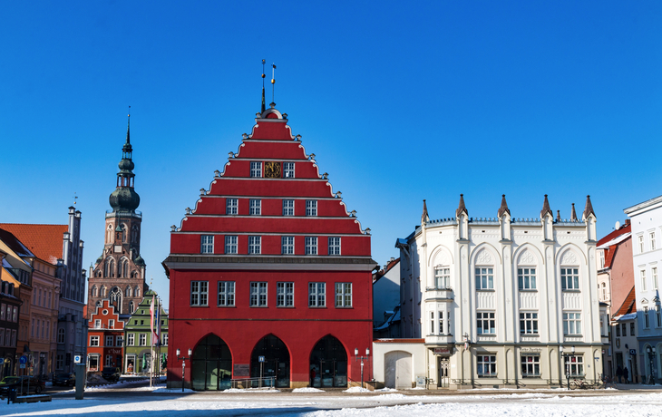 Historische Gebäude mit roter und weißer Fassade vor blauem Himmel und Kirche im Hintergrund. Greifswald