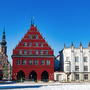 Historische Gebäude mit roter und weißer Fassade vor blauem Himmel und Kirche im Hintergrund. Greifswald