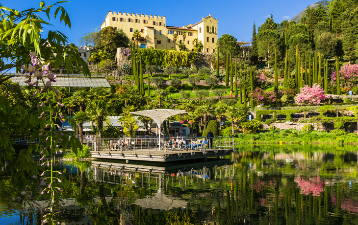 Gelbes Schloss inmitten grüner Gärten und Blüten, mit See im Vordergrund.
