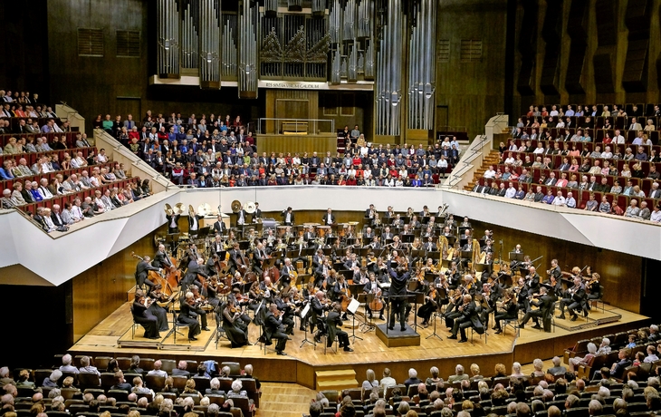 Ein Orchester spielt vor einem großen Publikum in einem Konzertsaal.