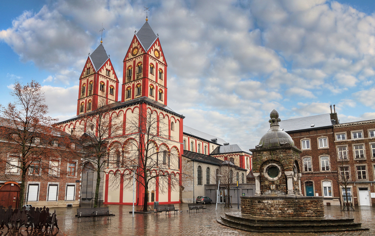 Kirche mit zwei Türmen und Brunnen auf einem Platz bei bewölktem Himmel.
