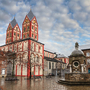 Kirche mit zwei Türmen und Brunnen auf einem Platz bei bewölktem Himmel.